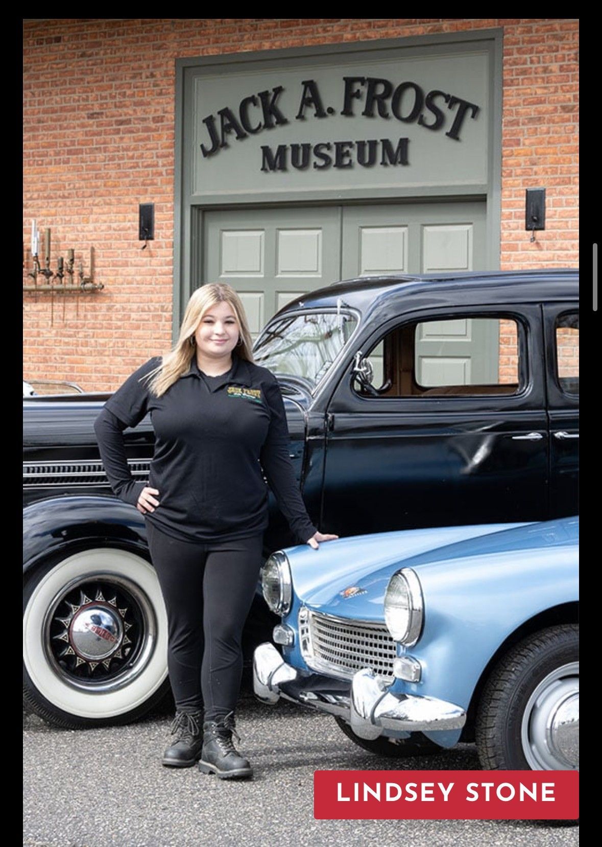 Woman stands by classic cars at the Jack A. Frost Museum, brick building in background.