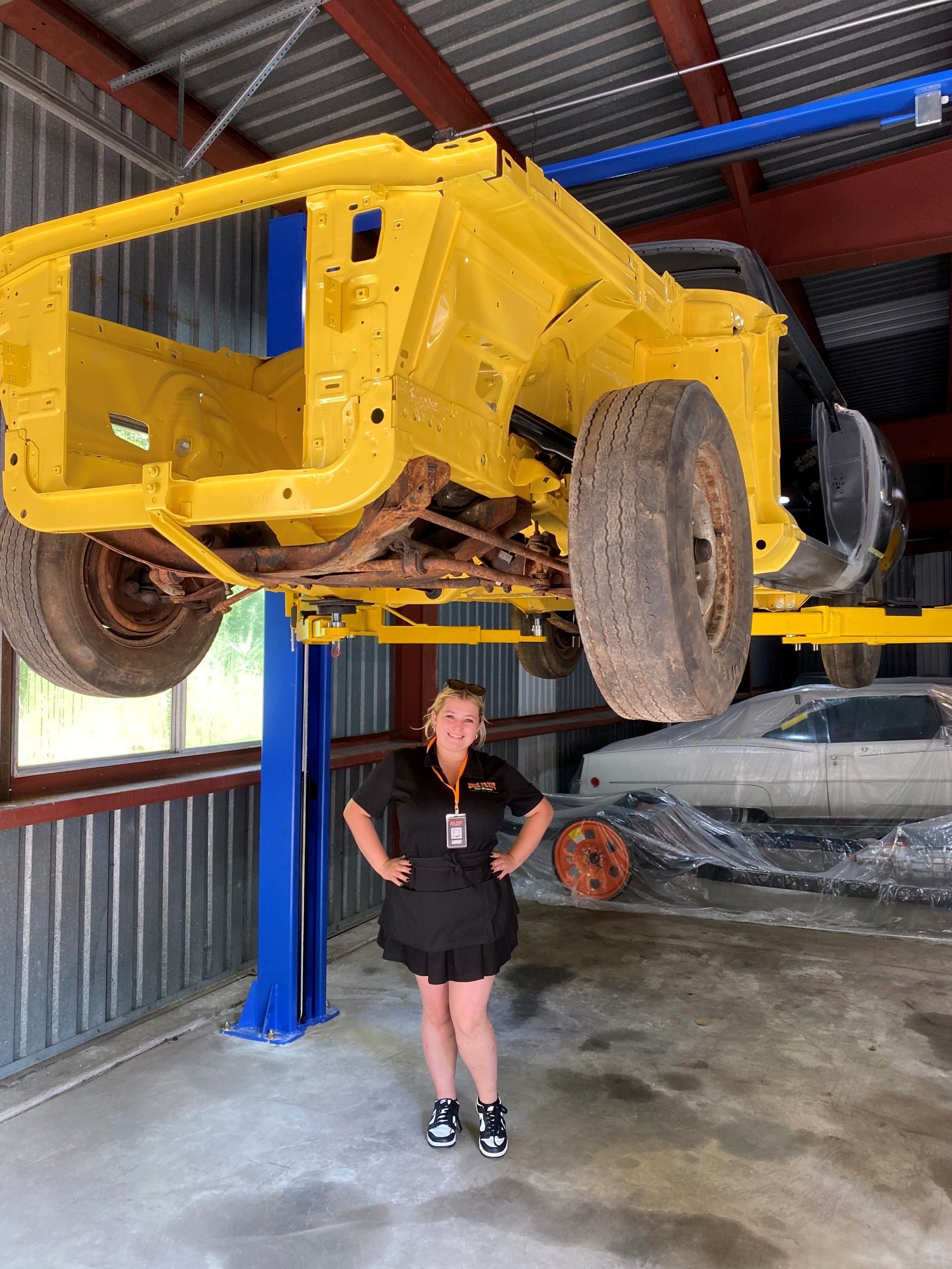 Woman in a black uniform stands beneath a yellow car chassis lifted on a hoist in a garage.