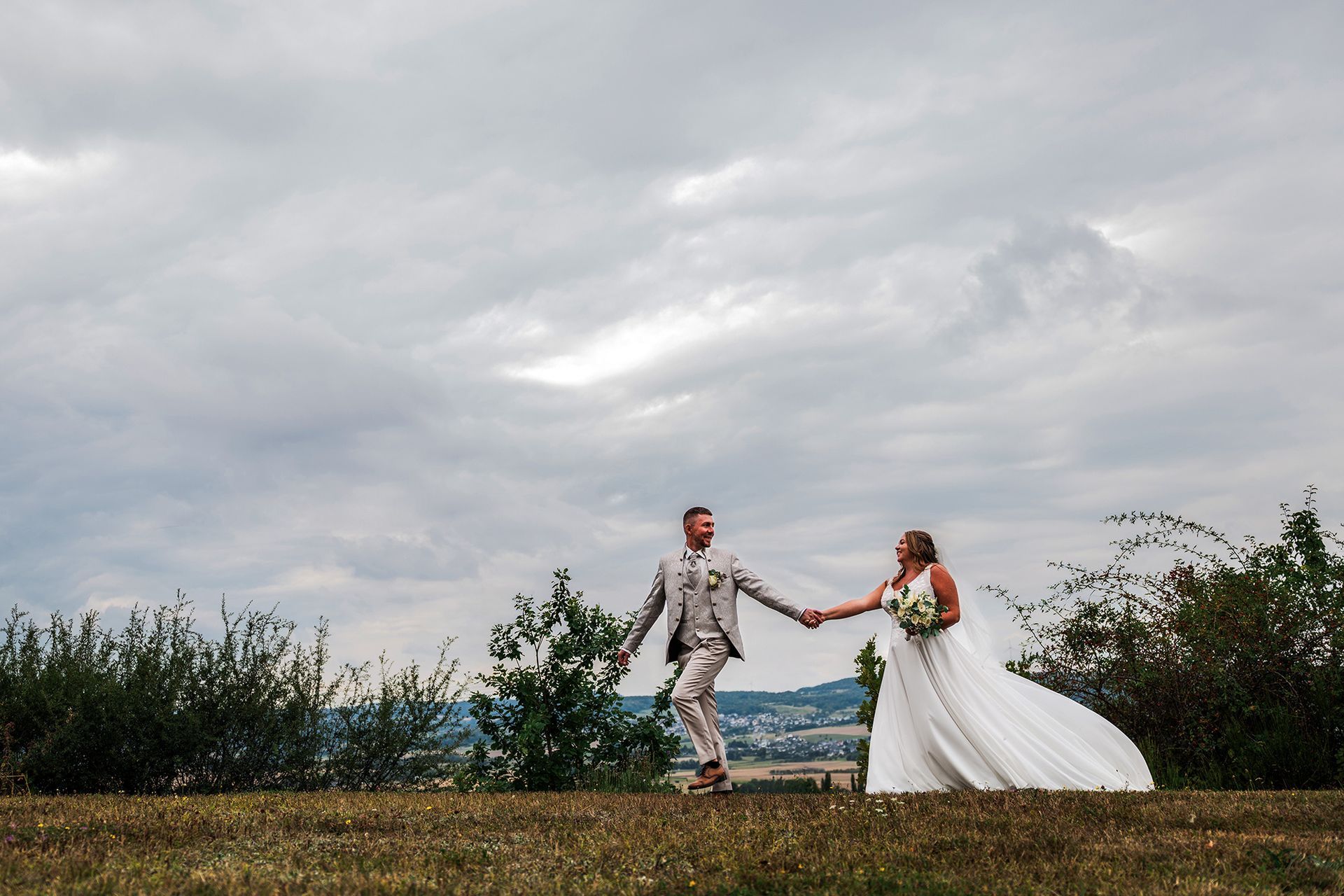 Ein Hochzeitsfoto mit laufendem Brautpaar. Das Kleid ist in voller Pracht zu sehen und weht im Wind.