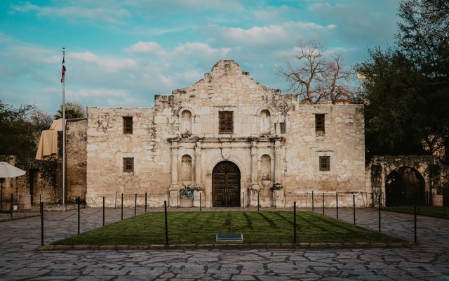 The Alamo mission in San Antonio, Texas. Stone facade with a central wooden door, under a blue sky.