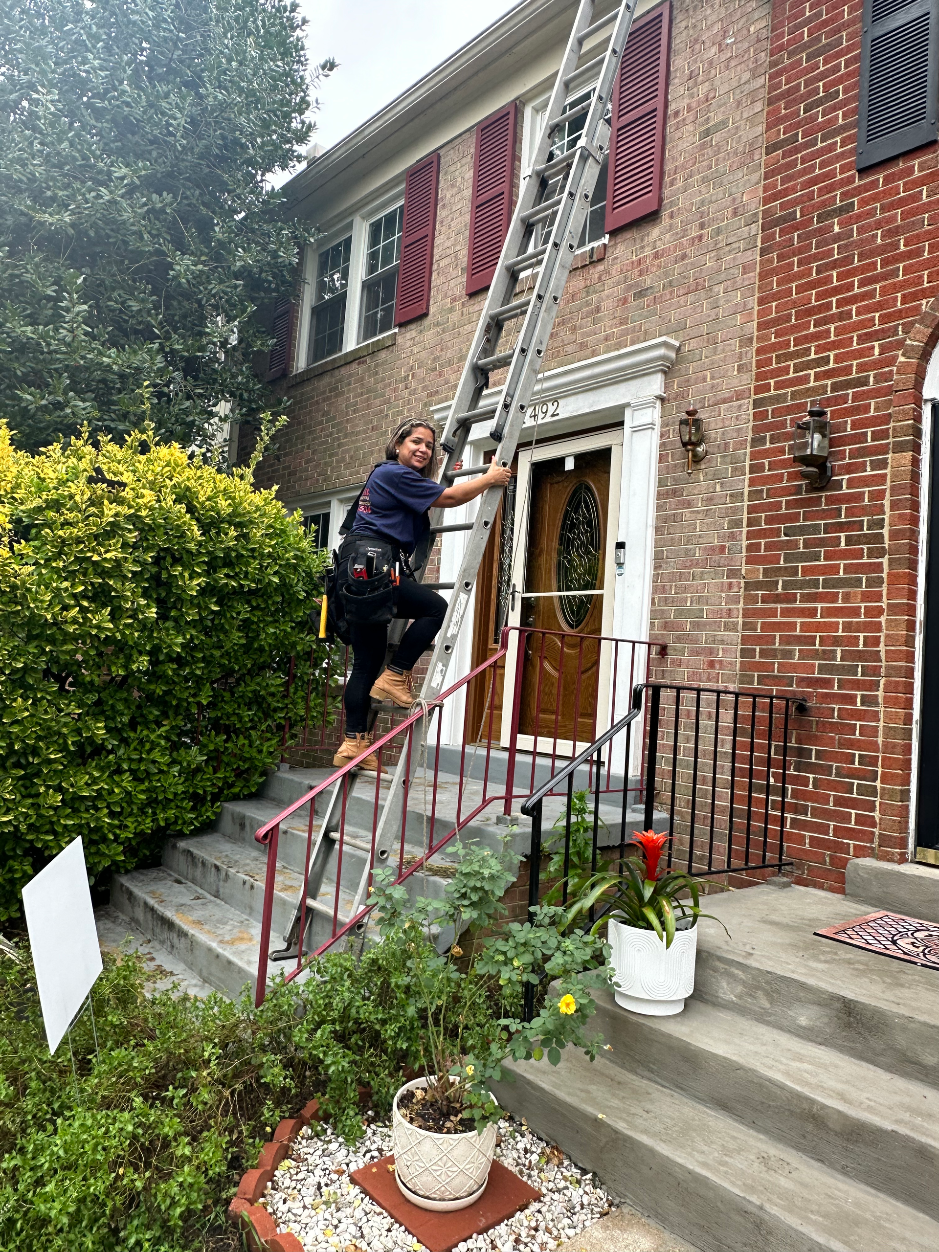 A woman is standing on a ladder in front of a brick house.