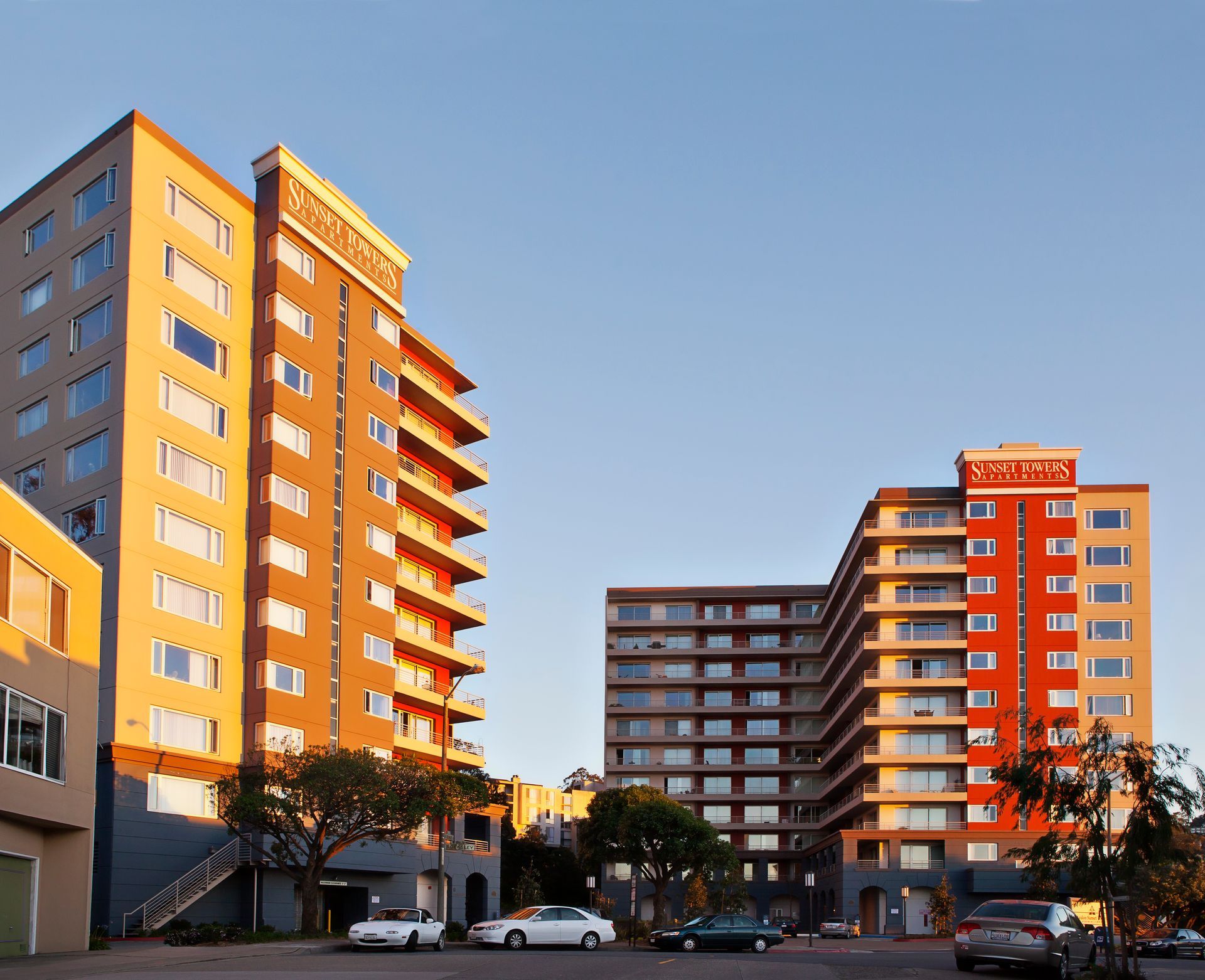 Two apartment buildings, one orange, one red, with cars parked in front under a clear sky.