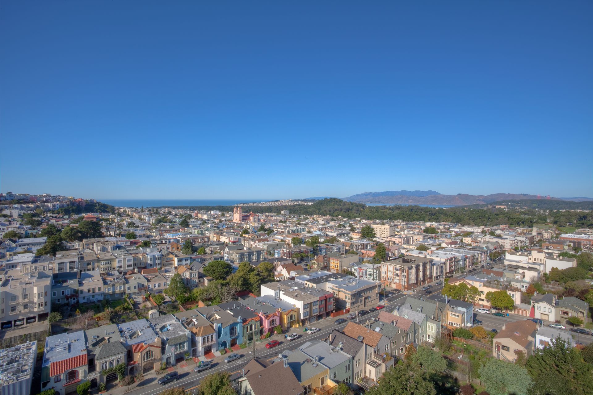 Cityscape with colorful houses under a clear blue sky, distant hills and ocean visible.