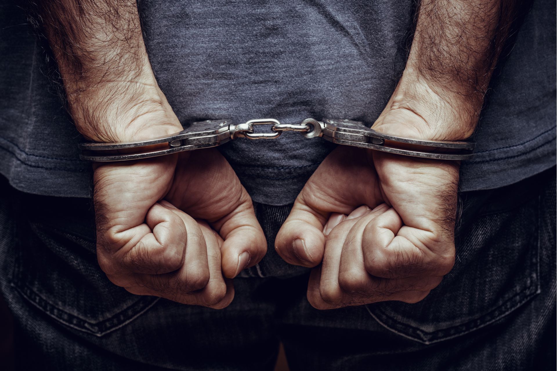 Person's hands cuffed behind back, wearing a gray shirt and jeans.