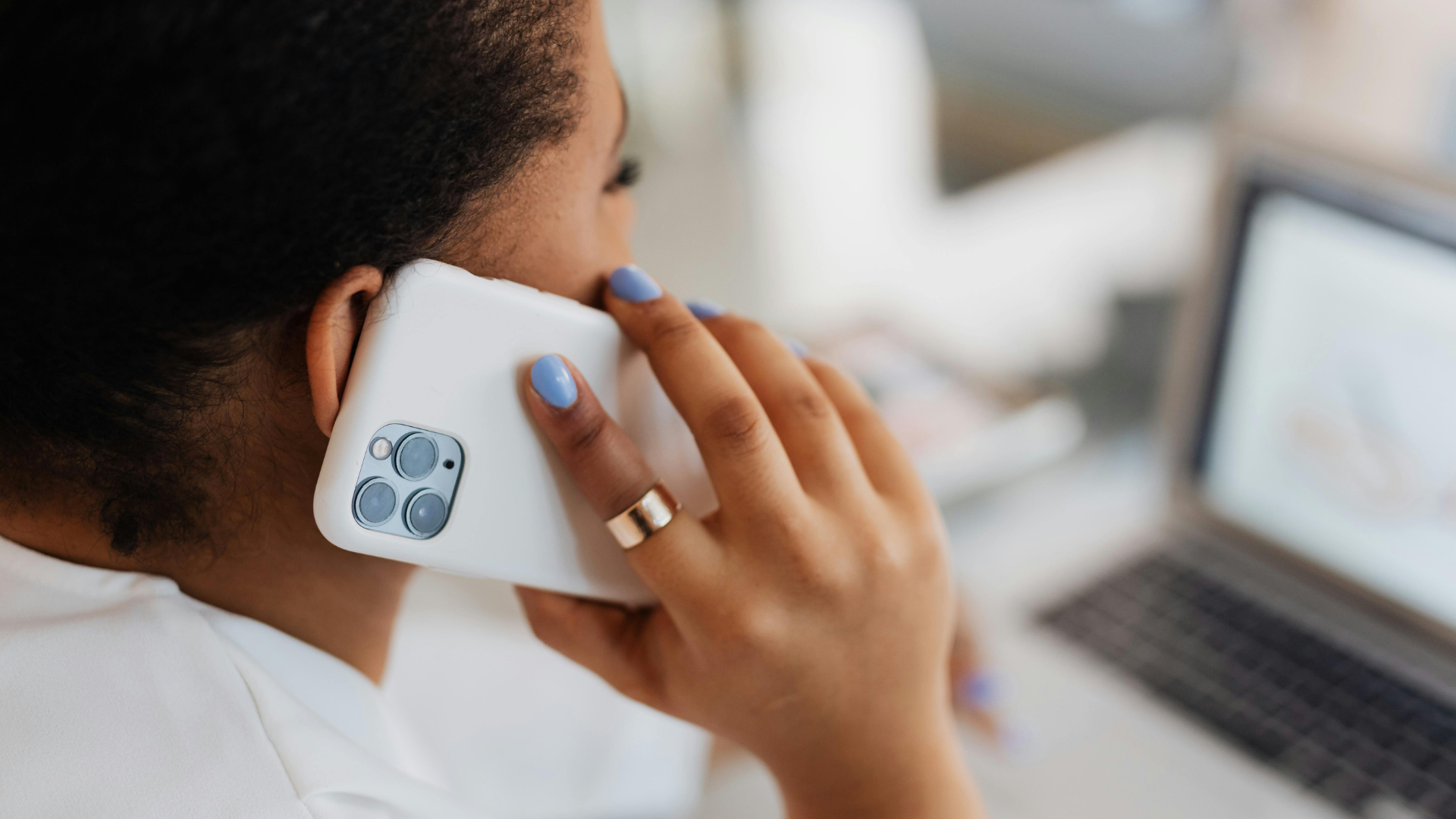 Person on a phone call with a laptop in the background. White phone, blue nails, gold ring.