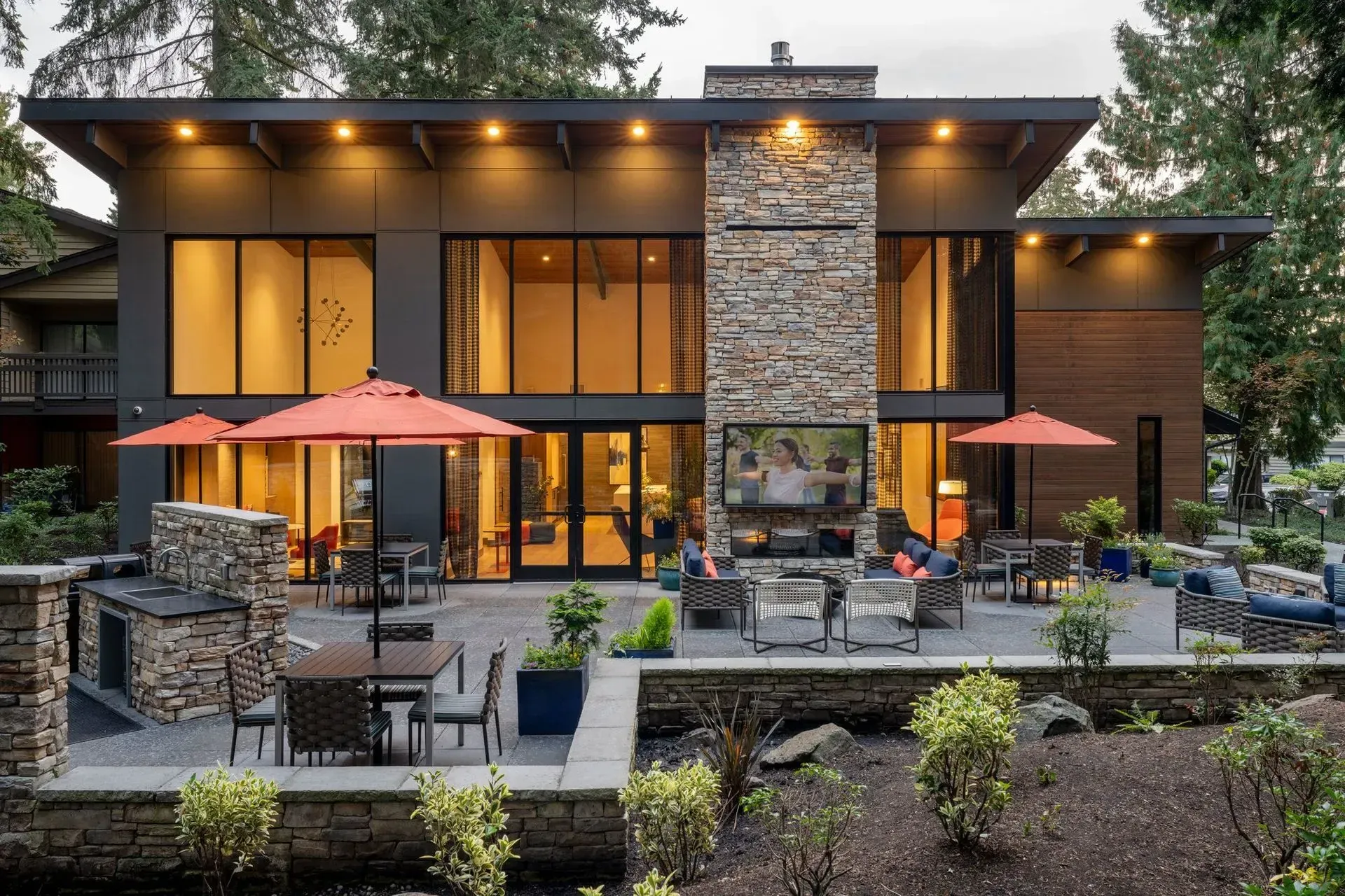 Apartment community outdoor amenities, including a resident lounge, grill station, and seating area with a TV, with the clubhouse building in the background.