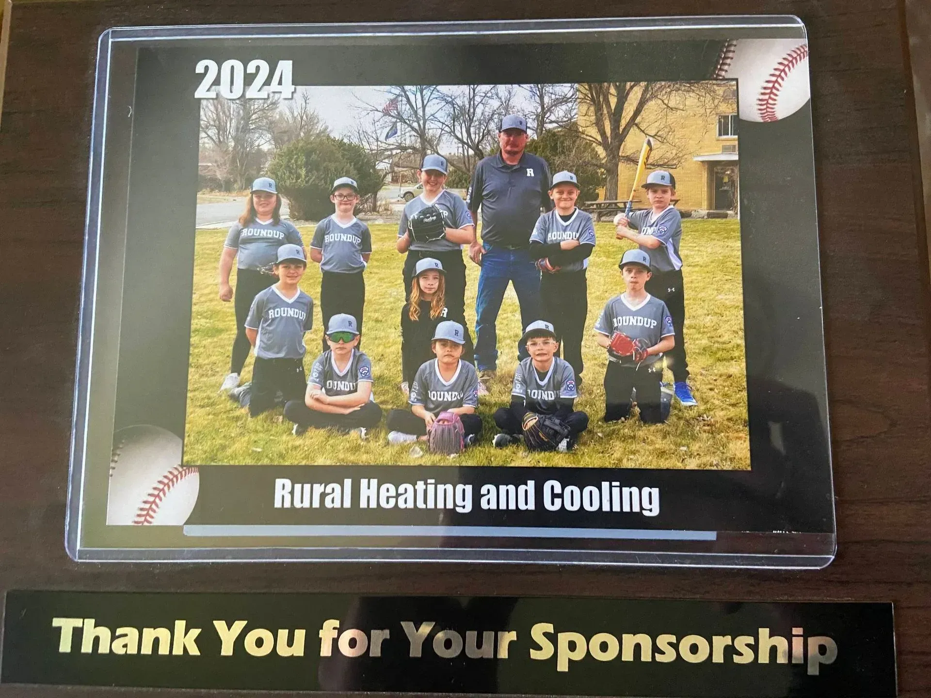 2024 youth baseball team photo with coach and sponsors. Team in gray uniforms, grass field, sponsor name at the bottom.