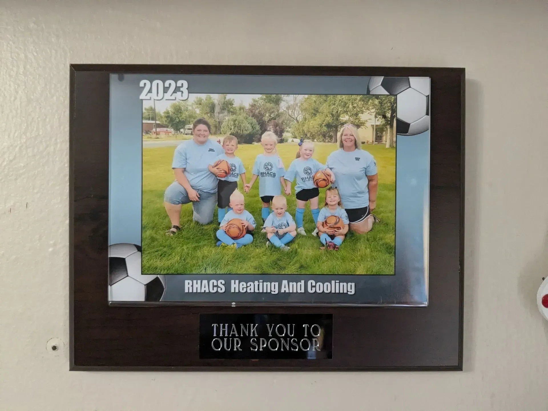 Team photo on plaque; soccer team with sponsor's logo. Dark wooden frame, 2023, soccer balls.