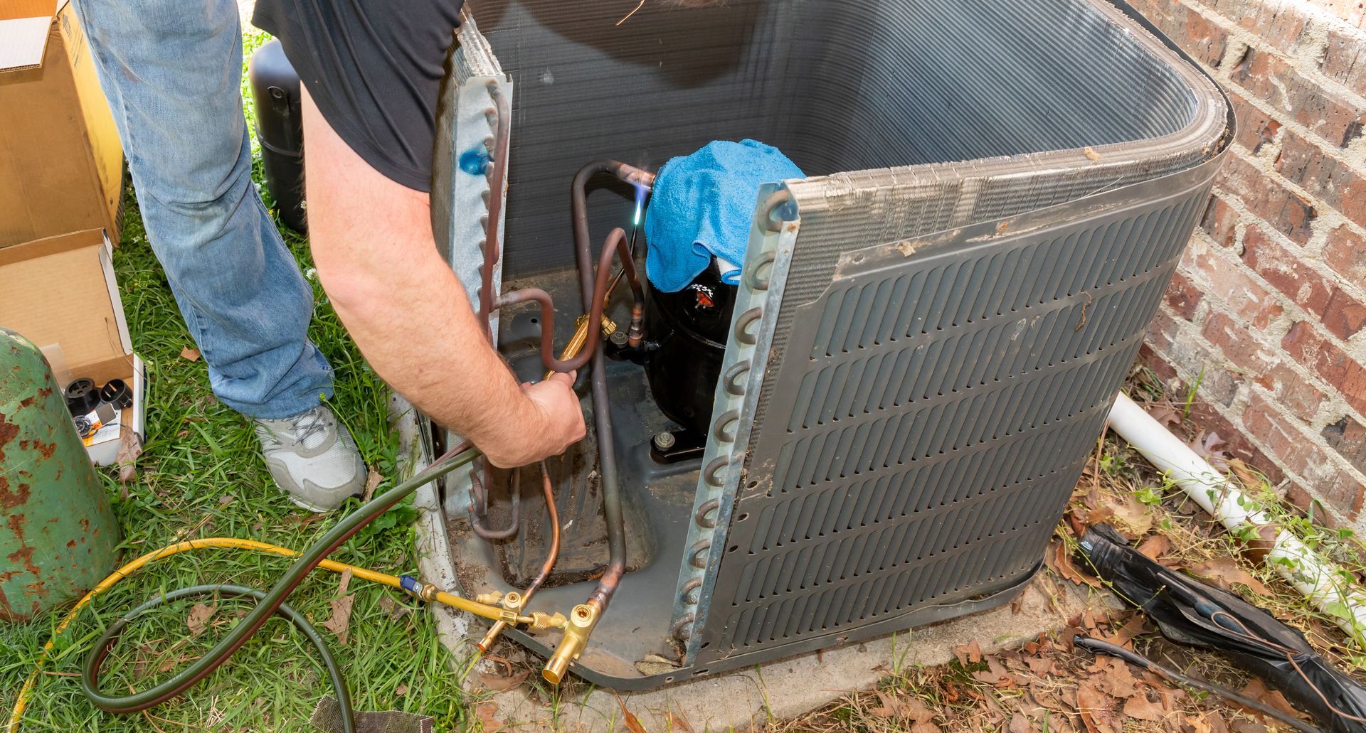A technician repairs an air conditioner outside; he connects lines to the compressor, near a brick wall.