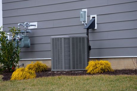 An air conditioning unit outside a house, with shrubs and gas meter visible.