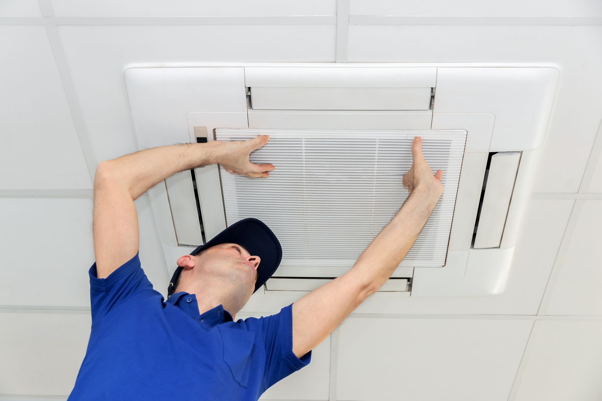 Man in blue shirt and cap installing an air conditioning filter in a white ceiling unit.