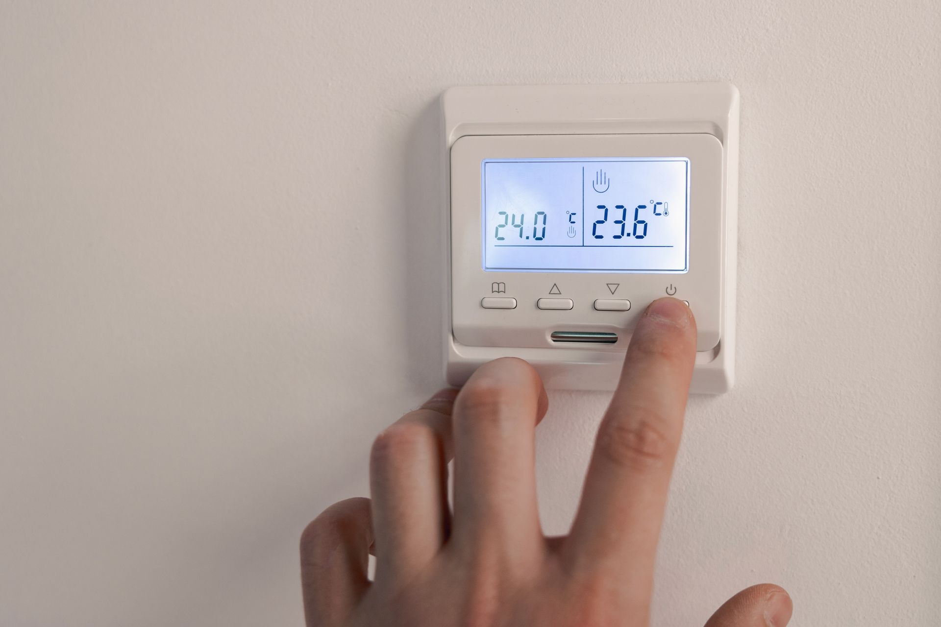 Person adjusting a white digital thermostat mounted on a white wall; the display shows temperatures.