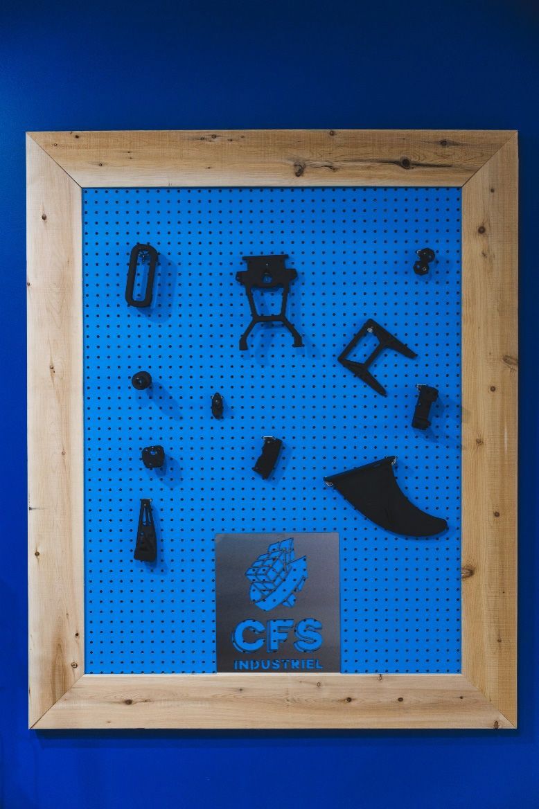 Objets noirs fixés sur un panneau perforé bleu à l'intérieur d'un cadre en bois, avec le logo « CFB » au centre.