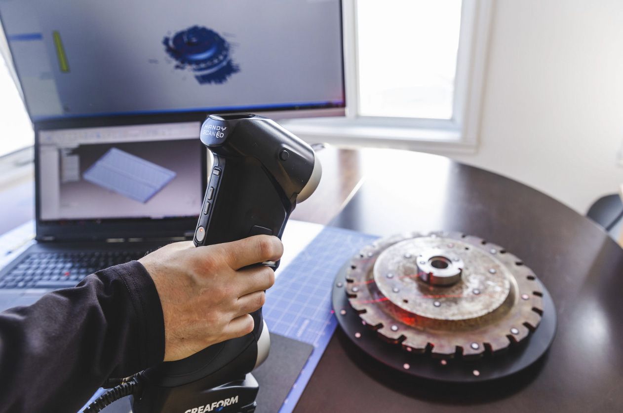 Une personne utilise un scanner portable pour numériser une pièce mécanique posée sur une table, un ordinateur portable affichant un rendu 3D.