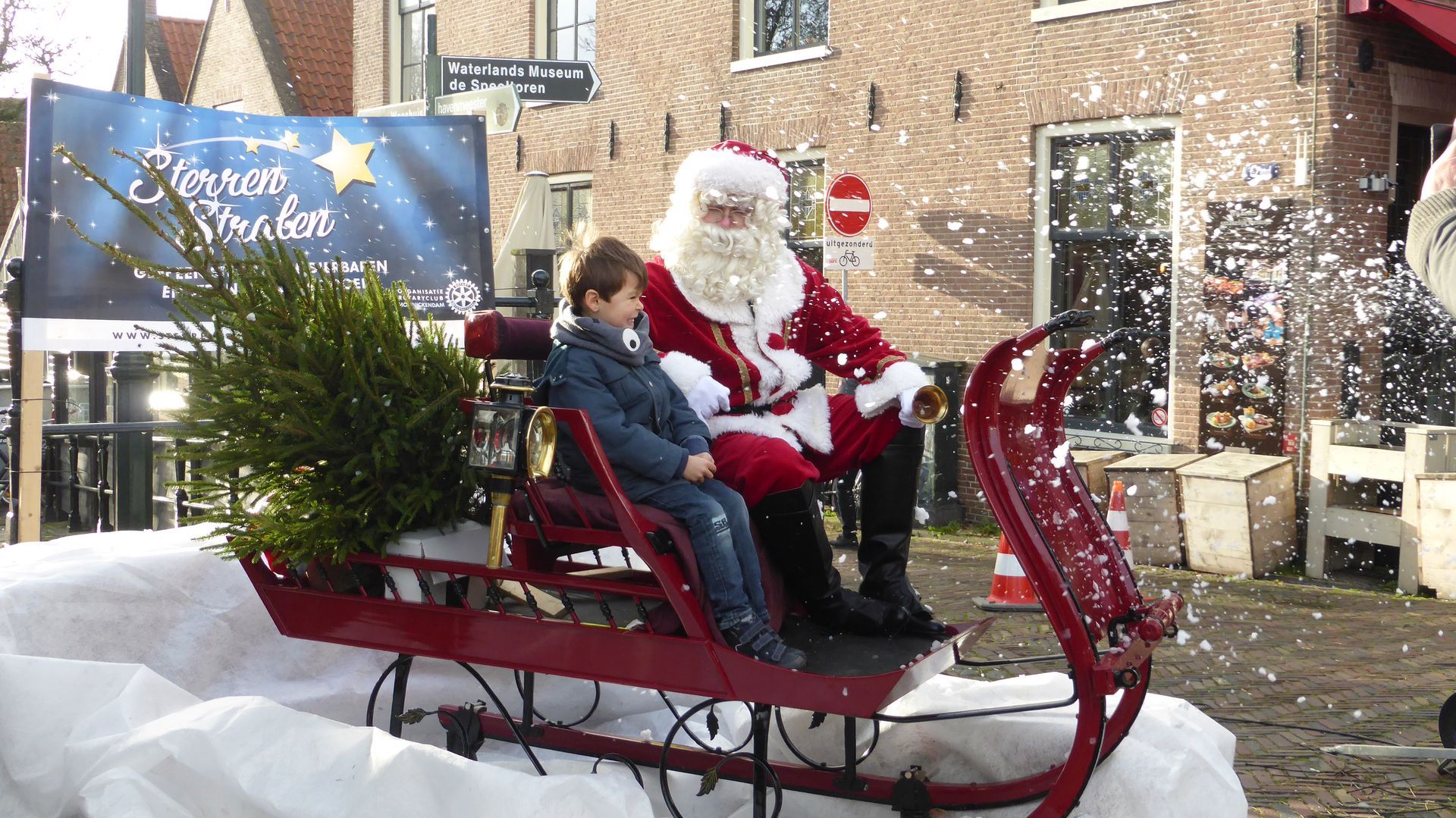 De Kerstman en een kind zitten in een rode slee met een kerstboom; er valt nepsneeuw.