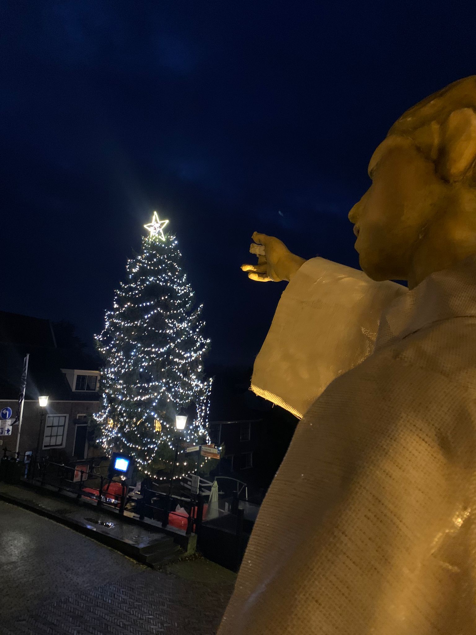 's Avonds wijst een beeld van een engel naar een verlichte kerstboom.