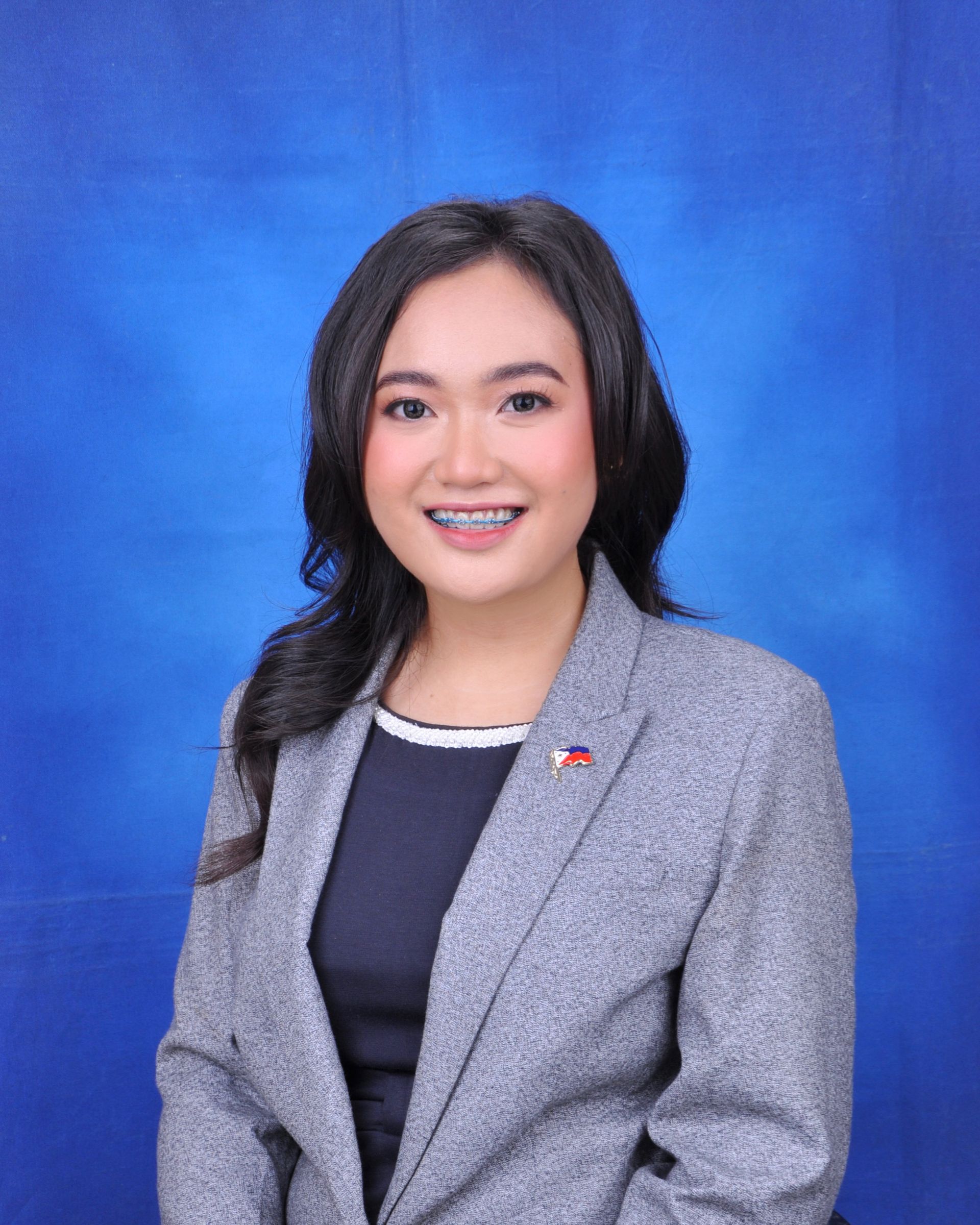 A woman wearing a gray jacket and a black shirt is standing in front of a blue background.