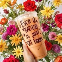 Hand holding iced coffee with “Even on bad days, just stay” surrounded by colorful flowers