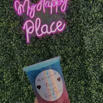 Hand holding a blue-capped snack cup in front of a neon pink “Mydappy Place” sign on green foliage wall