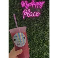 Pink drink cup held in front of a green wall with a pink neon “My happy place” sign