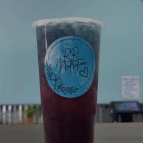 A clear cup of dark drink with a blue logo sticker on a beachside table.