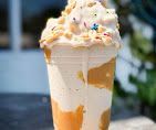Whipped cream-topped caramel milkshake in a clear plastic cup with sprinkles