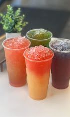 Four colorful iced drinks in plastic cups on a table, including red, green, and dark purple beverages.