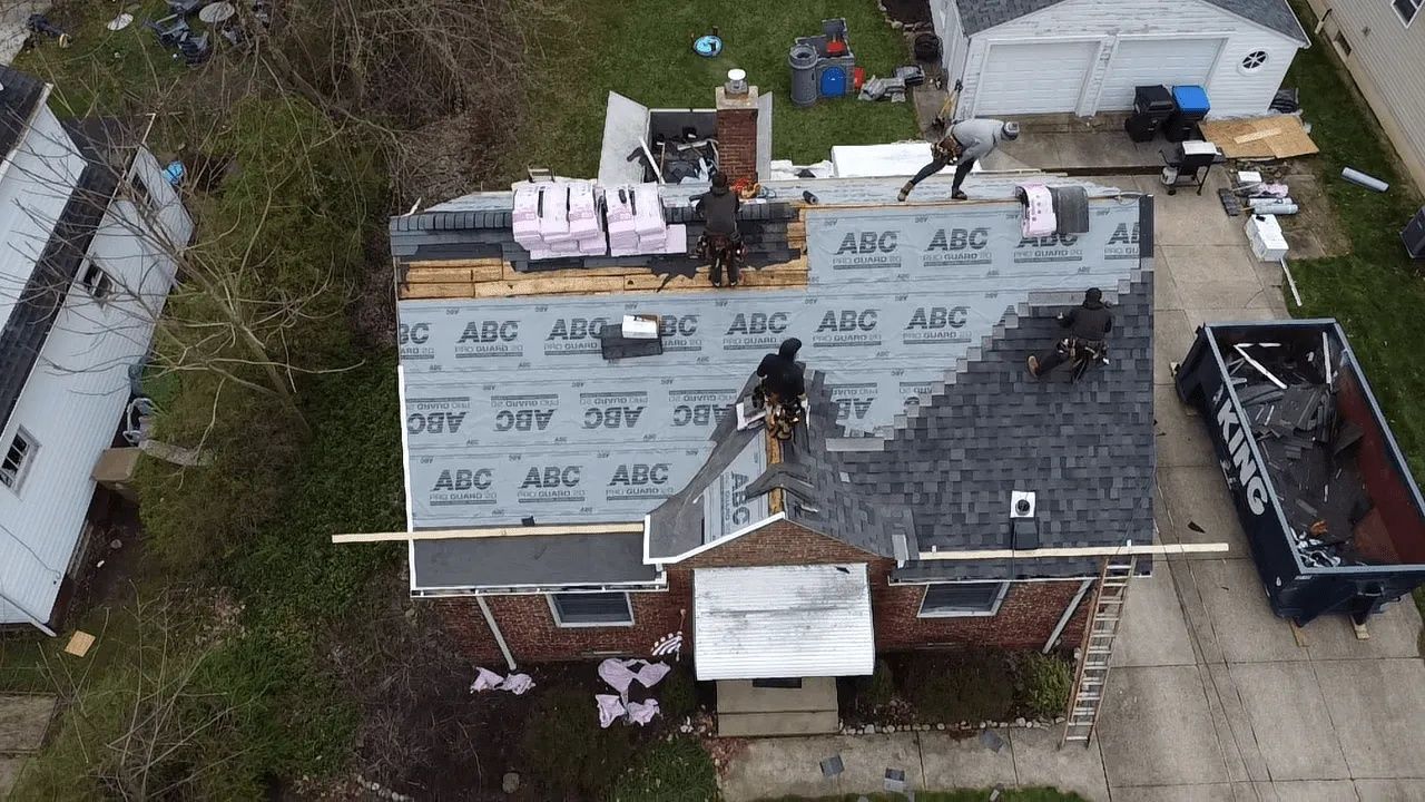 Roofers install shingles on a house roof, with some shingling completed.