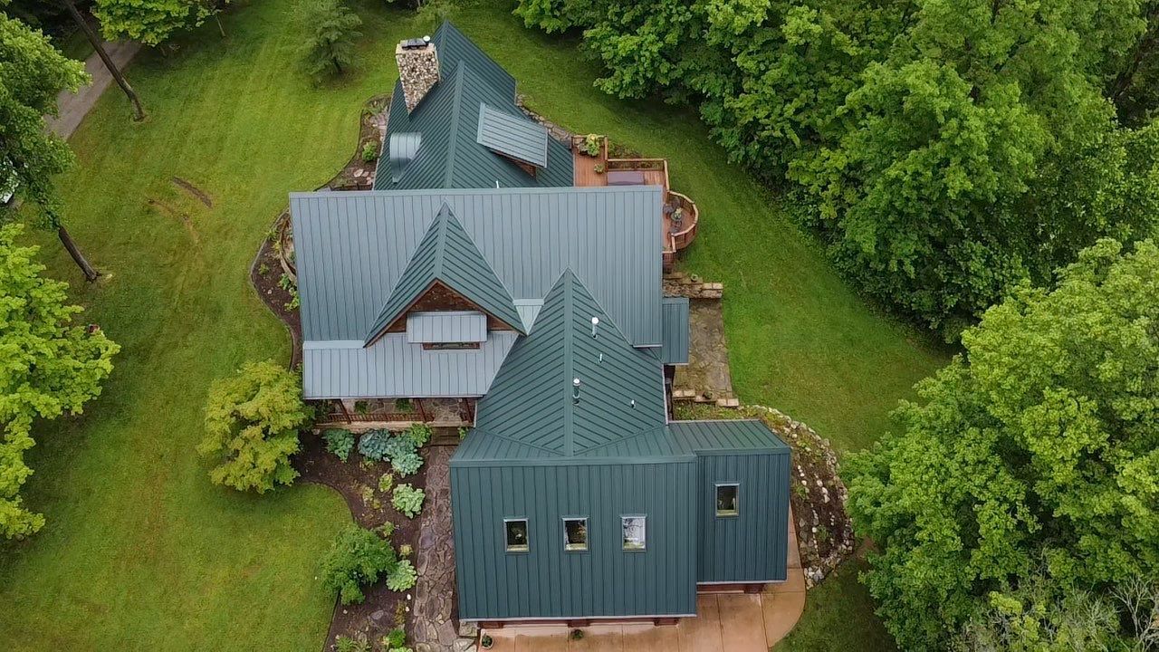 Overhead view of a house with a green metal roof, surrounded by green grass and trees.