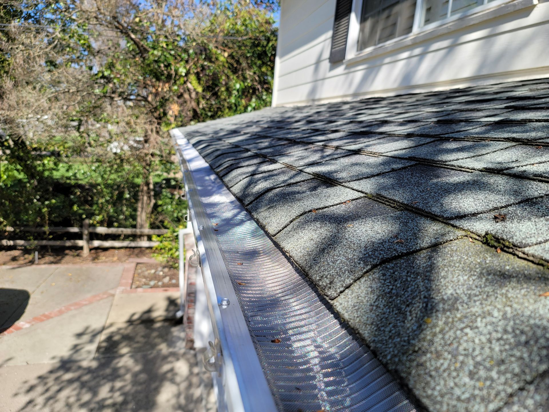 Gutter system with a mesh guard on a shingled roof, next to a white house with trees in the background.