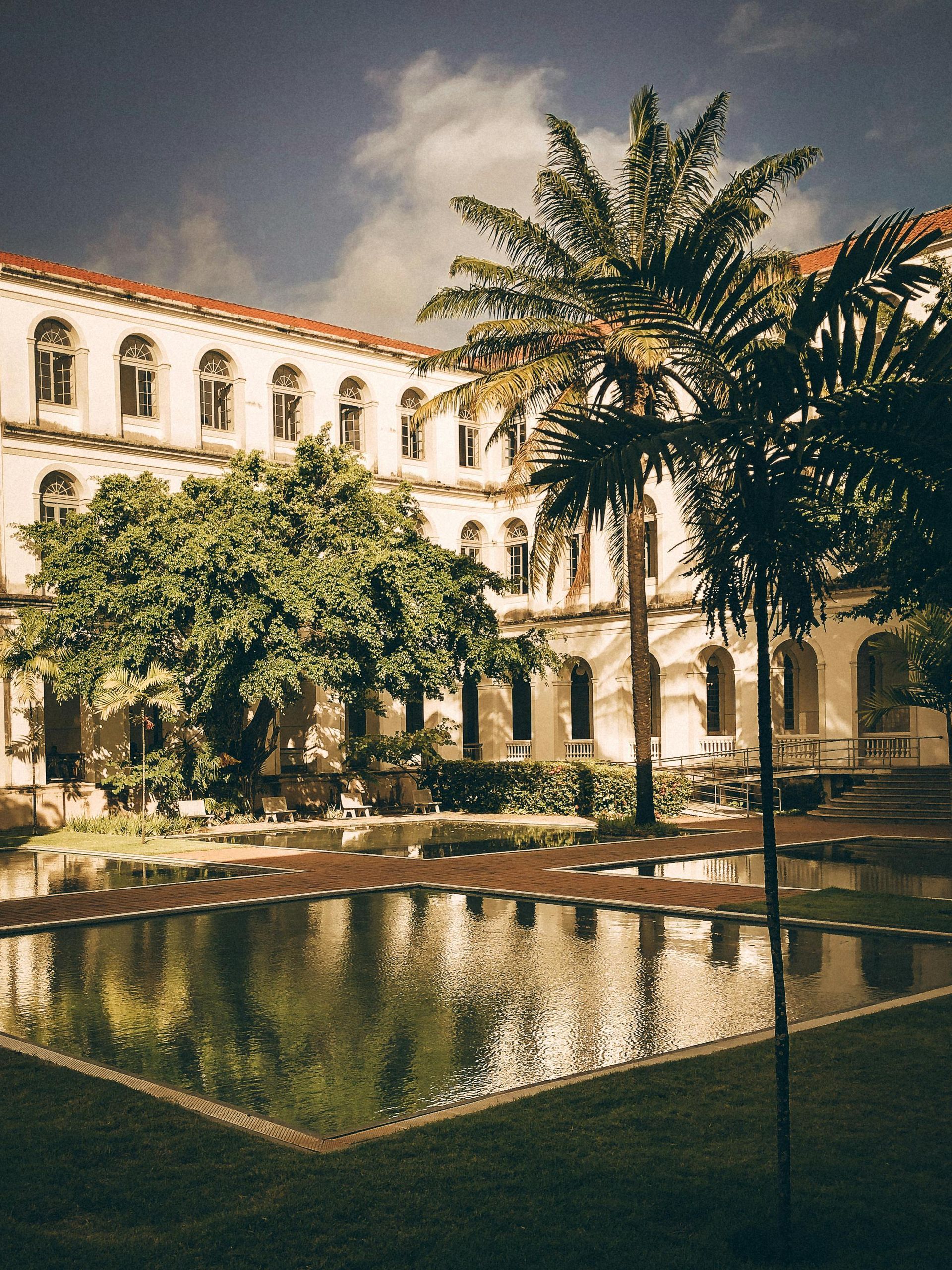A classic, white building with a pool in the foreground, palm trees, and a cloudy sky.