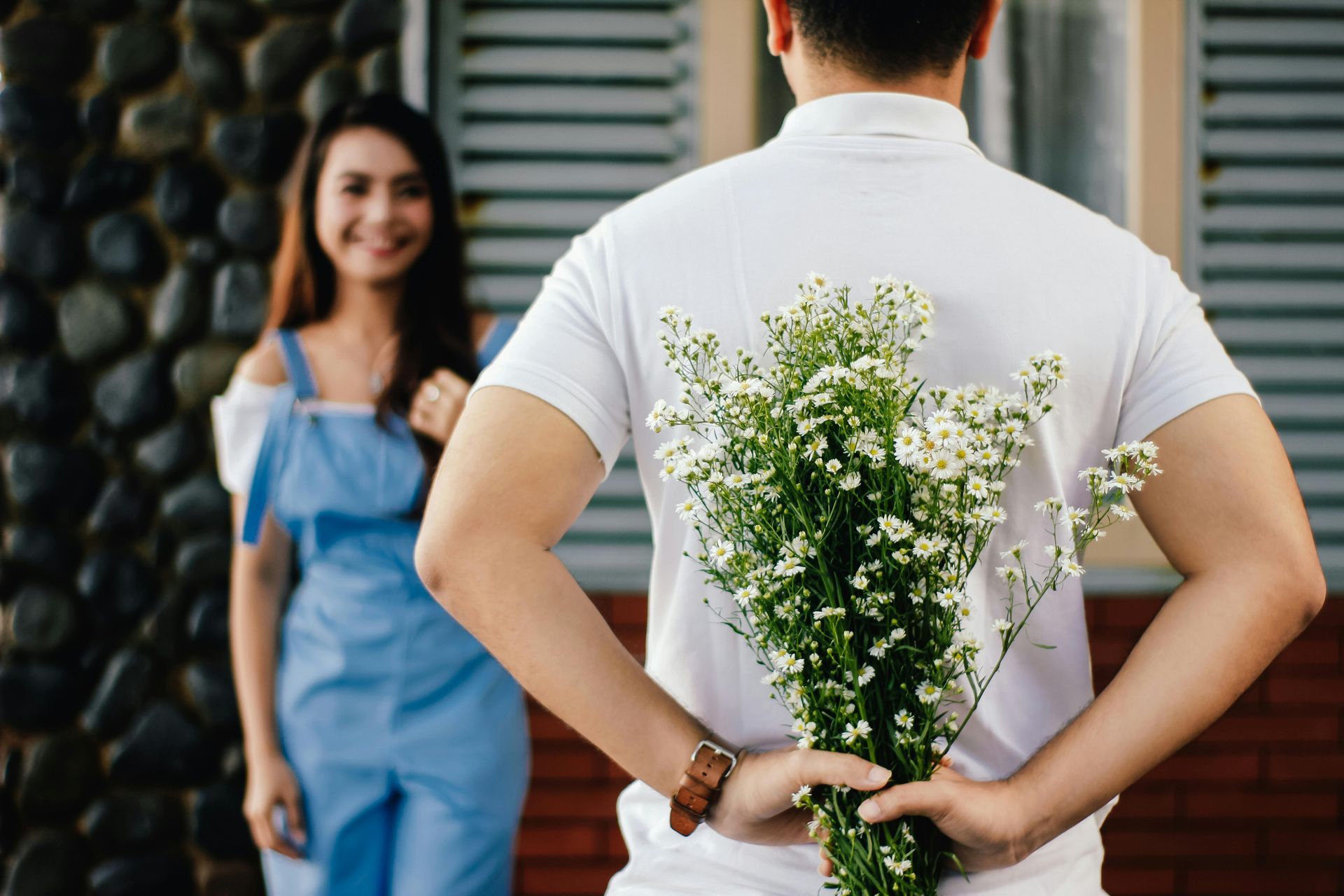 Man in white shirt holding flowers behind his back, surprising a smiling woman in blue overalls.