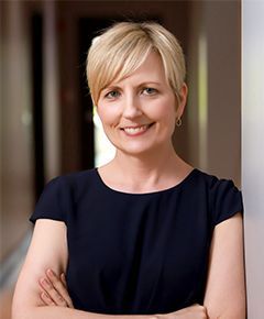 A woman in a black dress is leaning against a wall with her arms crossed and smiling.