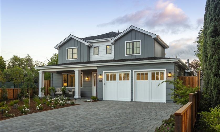 Two-story house with grey siding, white garage doors, and a paved driveway under a cloudy sky.