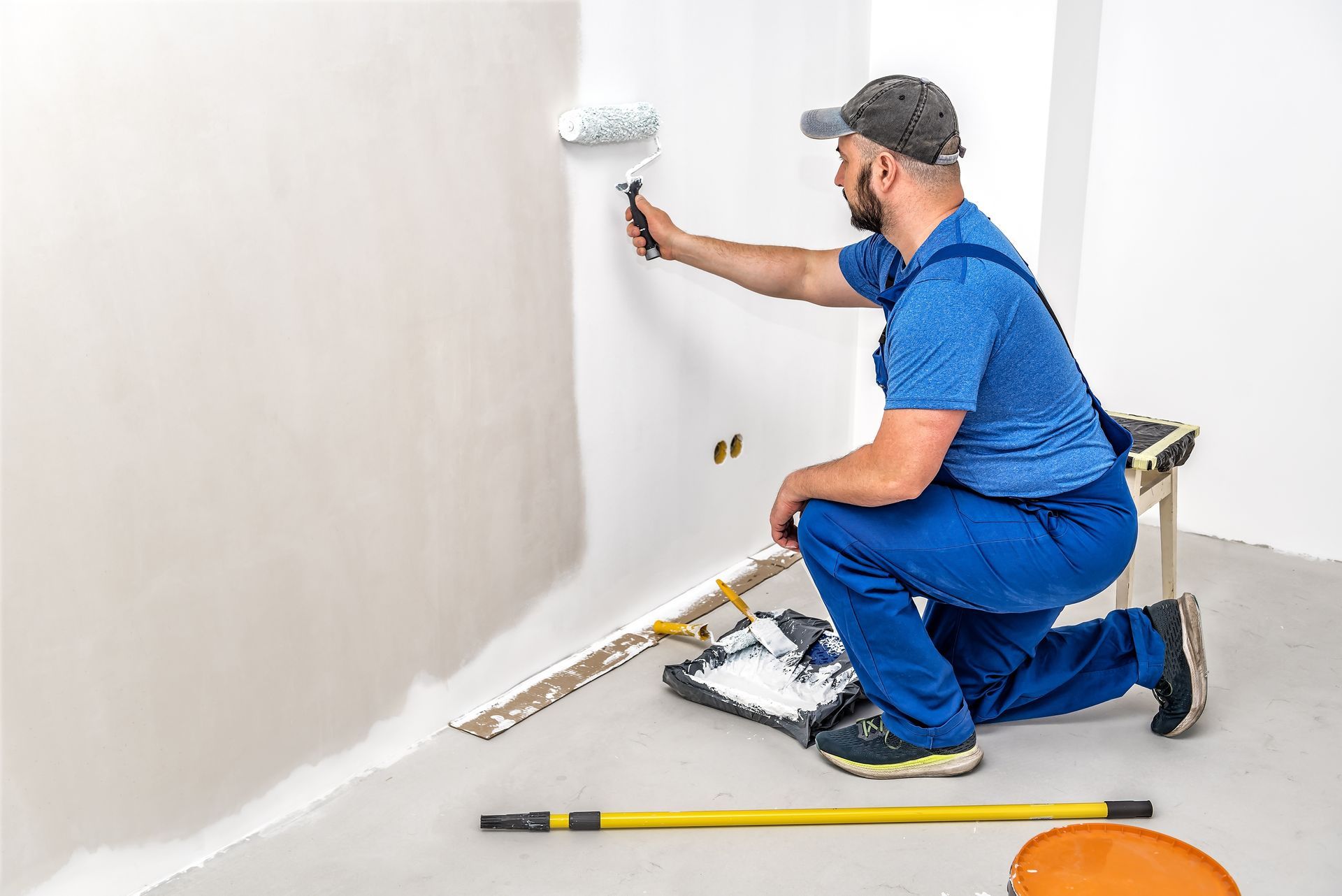 A man is kneeling down while painting a wall with a paint roller.