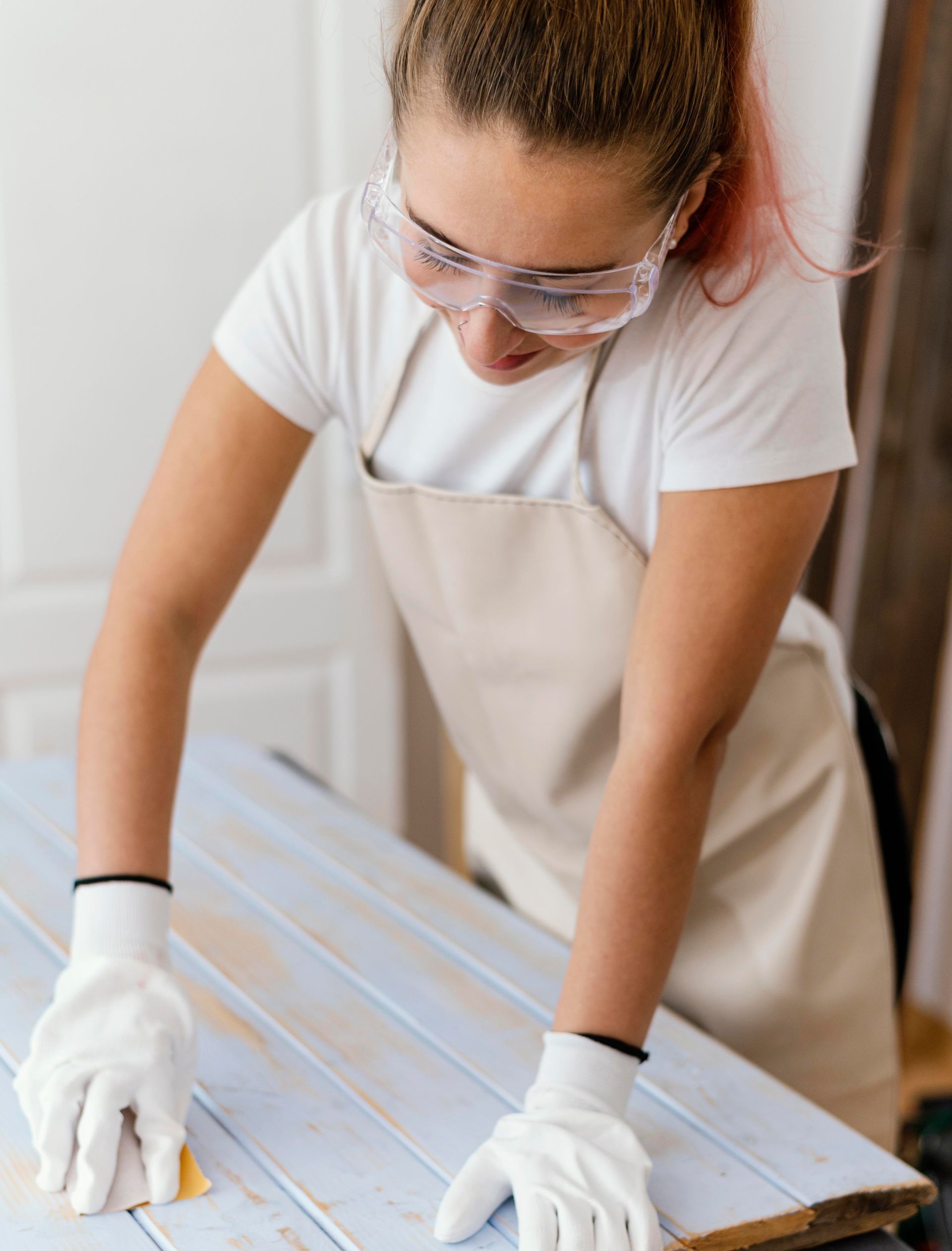 A woman wearing gloves and goggles is sanding a piece of wood.