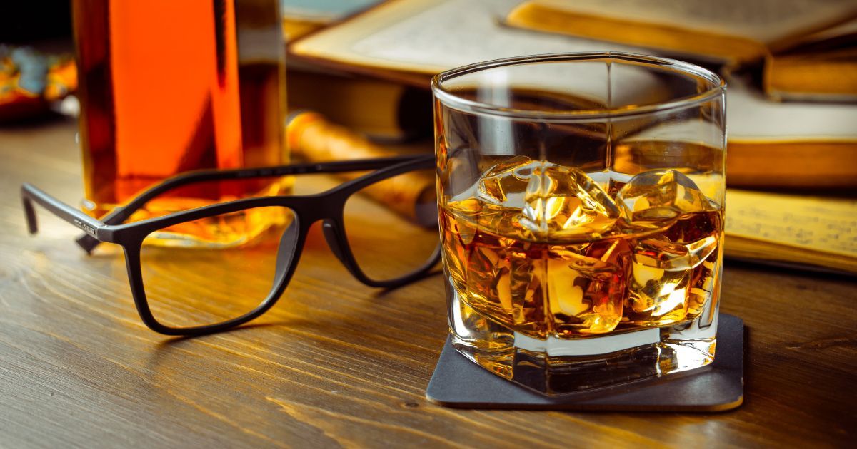 A glass of whiskey with ice cubes and a pair of glasses on a wooden table.