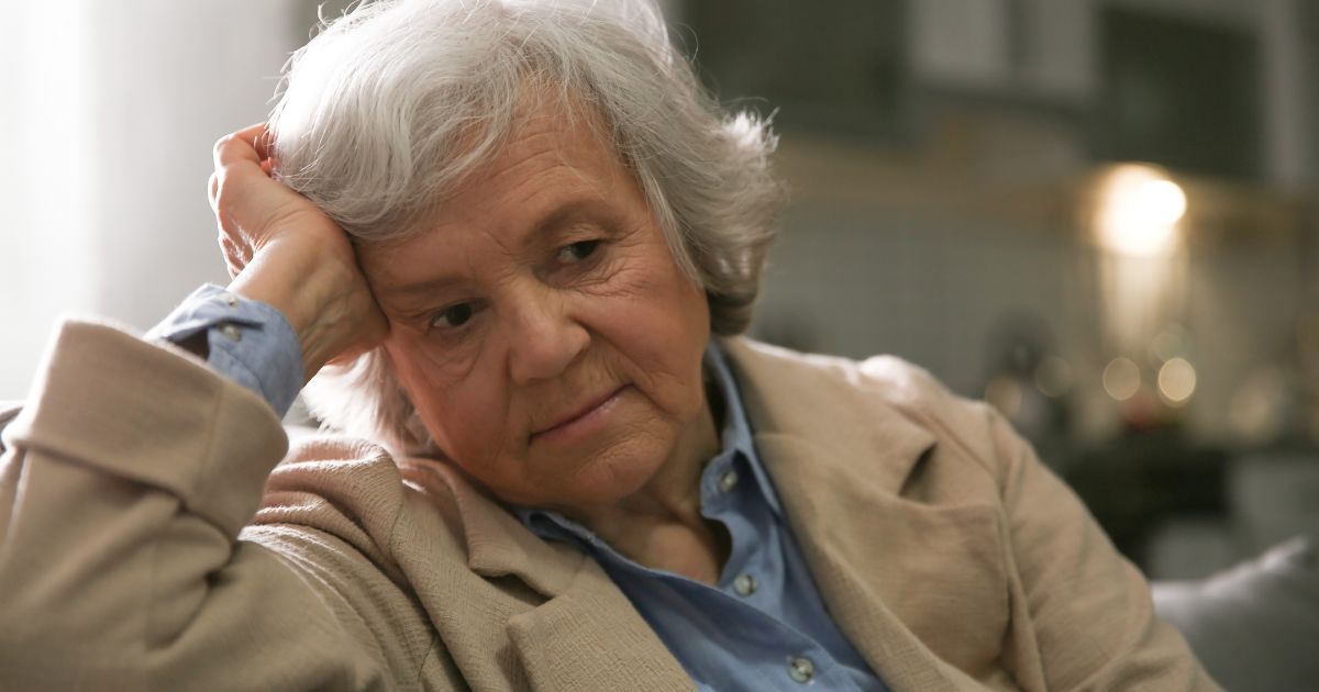 Older person with gray hair, looking down with a sad expression, resting head on hand.