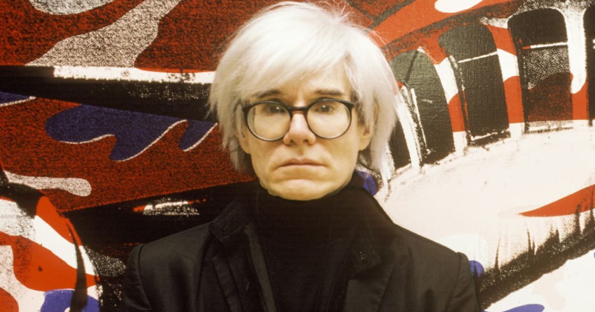 A man with white hair and glasses is standing in front of a painting.