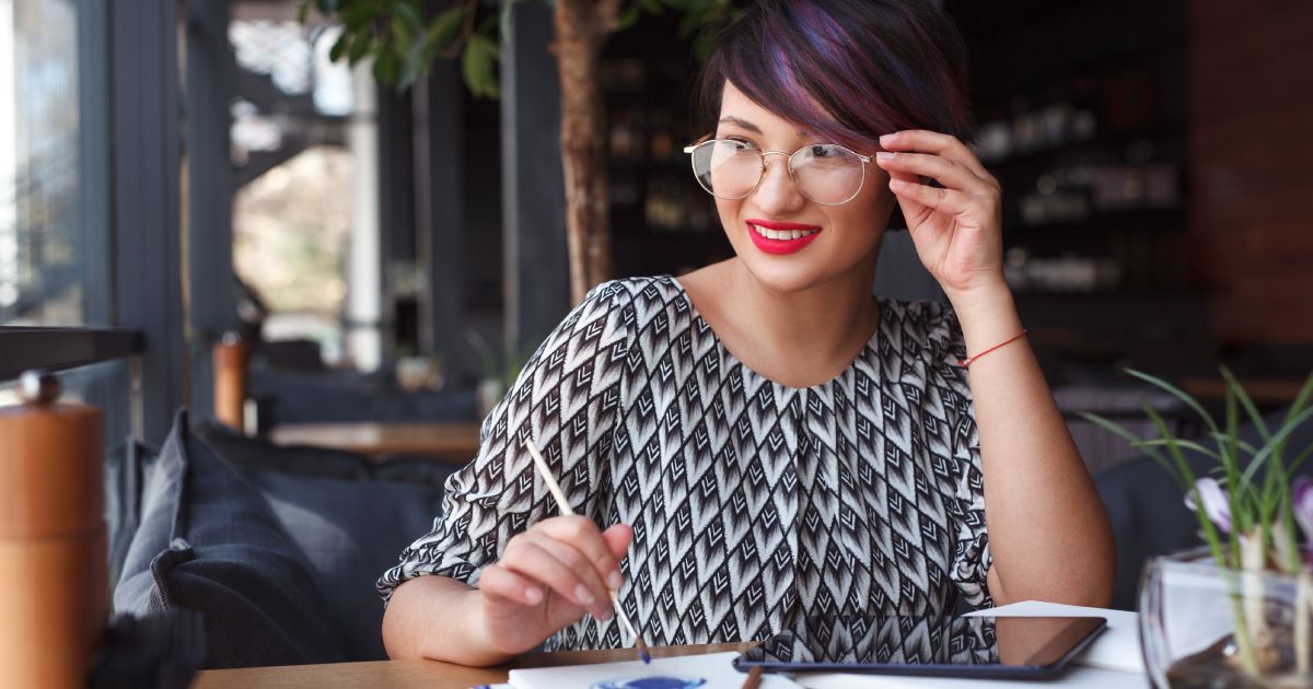 A woman in glasses  is sitting at a table with a tablet and a pen.