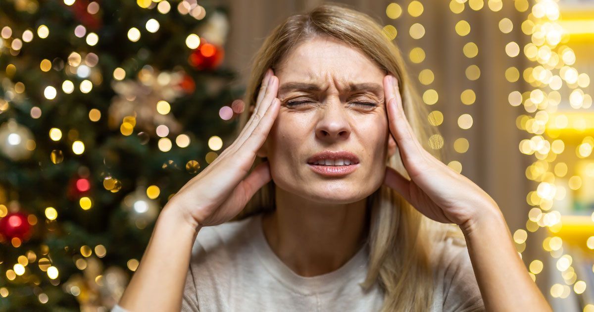 A woman sitting in front of a christmas tree is holding her hands to her head with her eyes closed.