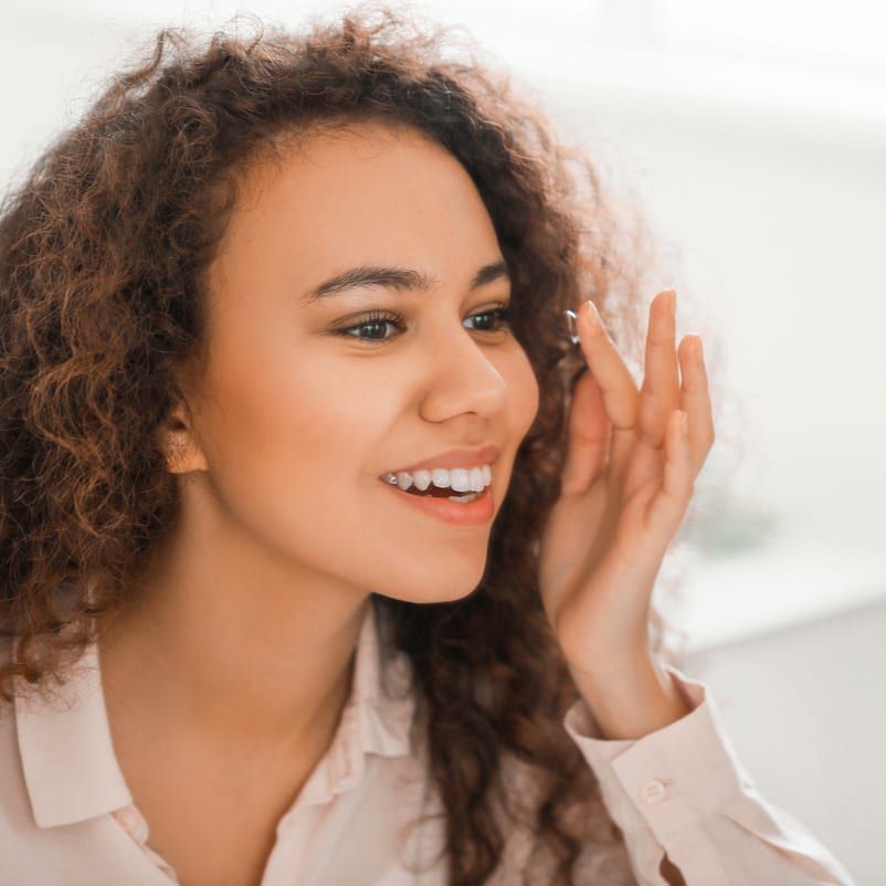 A woman with curly hair is putting a contact lens in her eye while smiling