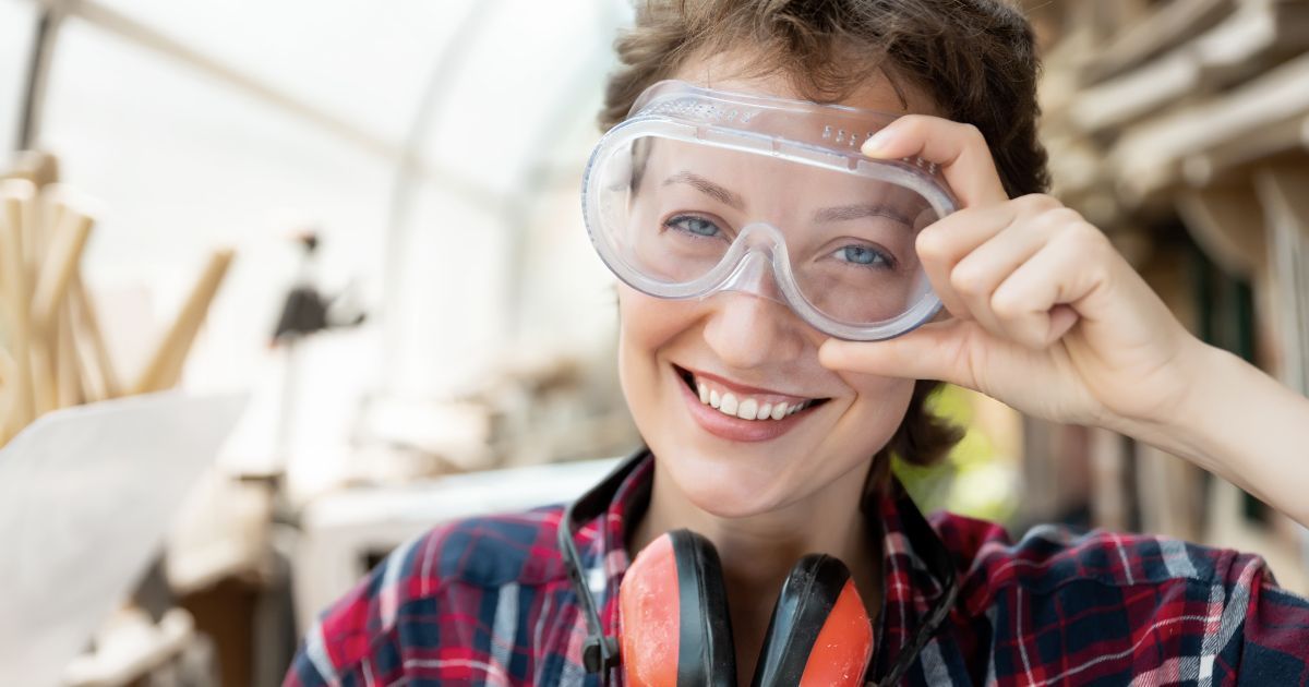 A woman wearing safety goggles and ear muffs is smiling.