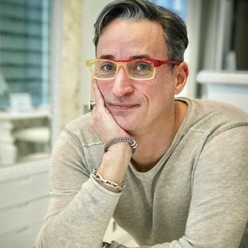 Designer Pierro Masarro  is wearing glasses while sitting at a table with his hand on his chin.