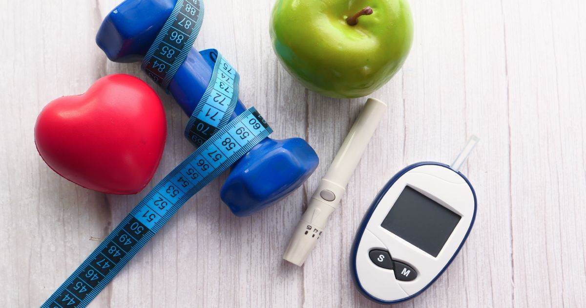 A diabetes monitor with a dumbell, an apple, a heart, and a measuring tape on a wood background.