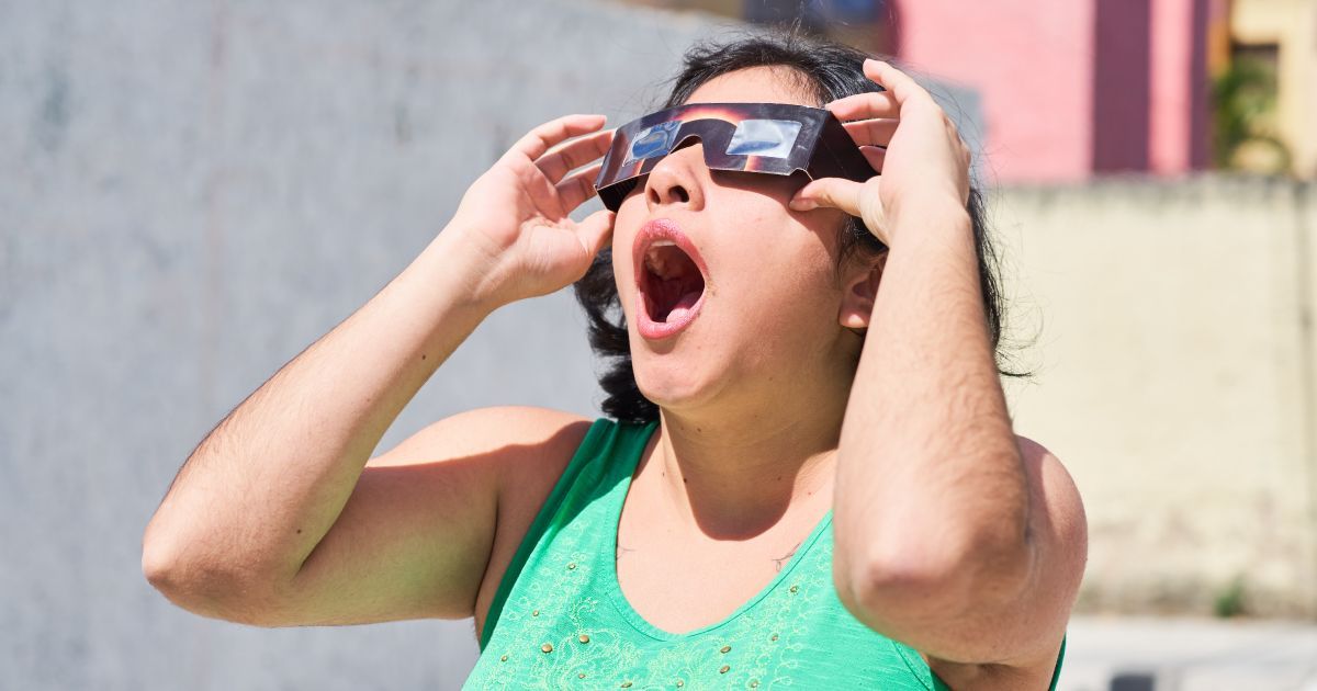 A woman wearing safety glasses looking up at a solar eclipse with her mouth open.