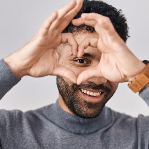 A man with a beard is making a heart shape with his hands over one eye