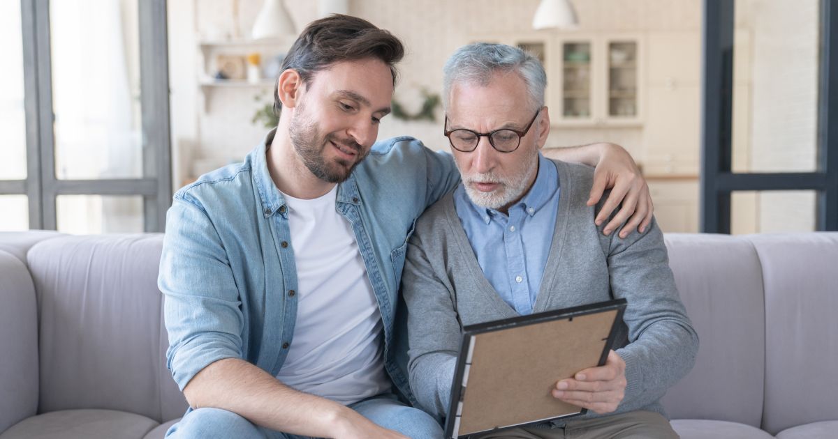 A son and a father are sitting on a couch looking at a framed photo.