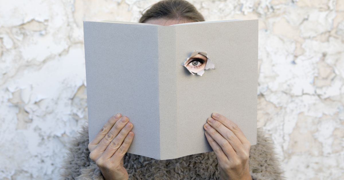 A woman is reading a book with a hole in the cover exposing her eye.