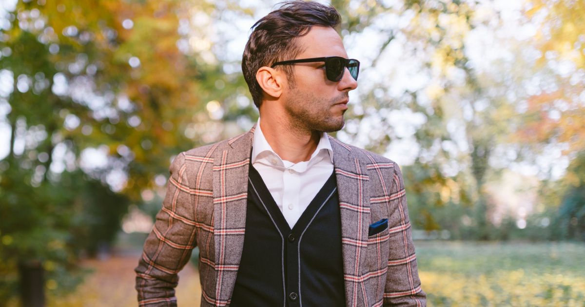 Man in plaid blazer and sunglasses, standing outdoors with autumn foliage.